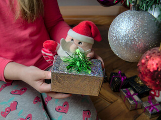 A girl sitting on the floor by the Christmas tree and holding a small Christmas present and a Santa Claus toy in her hands