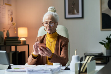Portrait of elderly woman sitting at office desk holding phone in hand. Wearing glasses and golden earrings, she is engaged with her phone and documents on her desk