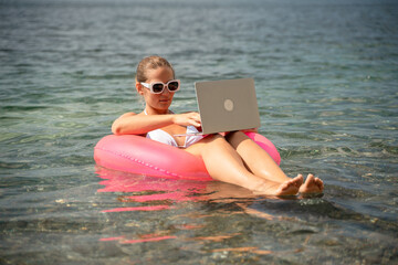 A woman is sitting on a pink inflatable raft in the ocean, using a laptop. Concept of relaxation...