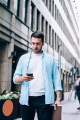 Serious man reading received message about smartphone notification while installing banking application for making online money transfer, Caucasian hipster guy standing at urban setting with cellular