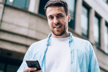 Half length portrait of positive male blogger standing outdoors and smiling at camera during time...