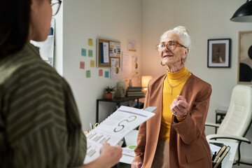Elderly businesswoman conversing with coworker in office filled with charts and posters. Engaging...