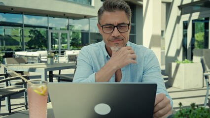 Focused businessman working on laptop on modern office terrace. Happy mid adult, mature man in glasses, deep in work, thinking, solving business problem, making decision online.