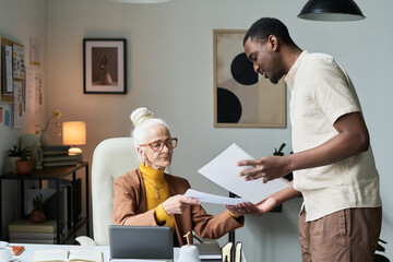 Obraz premium Elderly woman and young man engaged in business discussion in modern office setting, with papers, laptop, and plants around them. Background has creative elements and professional ambiance