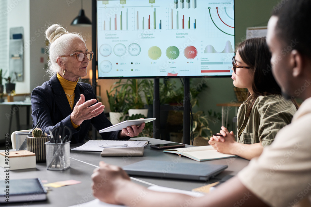 Poster Senior businesswoman presenting statistics using tablet to diverse team in cozy office setting with graphs displayed on large screen in background