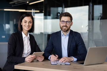 Business professionals collaborating on digital tablet and documents in modern office. Man and woman engaged in project discussion, showcasing teamwork and leadership skills with technology