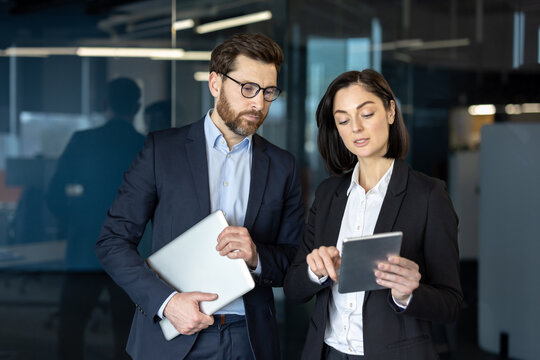 Business professionals engaged in discussion using tablet in office setting. Man holding laptop while woman demonstrates with tablet, showcasing teamwork and communication in corporate environment