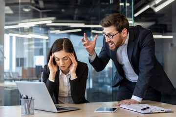 Angry boss yelling at stressed female office worker seated at desk with laptop and phone. Intense...