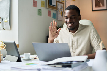 Friendly man using laptop while waving and smiling during virtual call in office setting. Workspace with sticky notes and decor visible in background