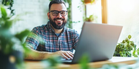 A bearded man joyfully works on his laptop in a bright space filled with lively green plants, embracing productivity and positivity in a natural setting.