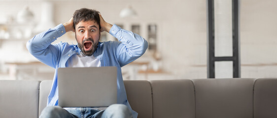 A man sitting on a couch at home is holding his head in frustration while staring at his laptop...