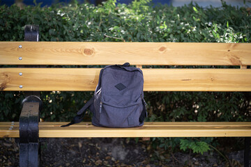 A grey backpack on a wooden bench in a park against a backdrop of green bushes.