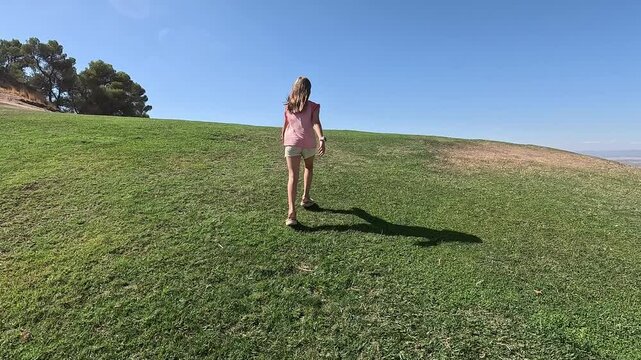 Following a preteen girl walking through a meadow of green grass with a horizon and blue sky and a huge plain full of olive crops is revealed