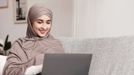 Freelance Concept. Young muslim woman in hijab working on laptop at home, sitting on couch, typing on keyboard and smiling