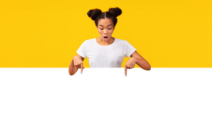 Amazed afro girl looking and pointing down to white blank placard ready for your text or banner. Empty space © Prostock-studio