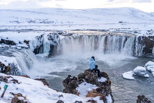 Woman hiker sitting looking at frozen and snowy Godafoss waterfall in winter