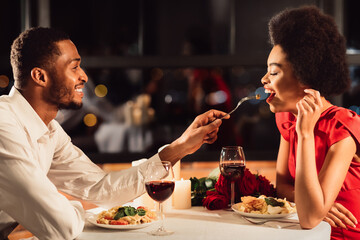 Romantic Date Concept. African American Man Feeding Girlfriend During Dinner Celebrating Anniversary In Restaurant
