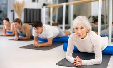 Slender elderly woman practicing pilates with blue roller in training area during pilates classes