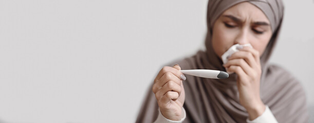 Seasonal Flu. Sick arabic woman in headscarf holding thermometer, checking her temperature and...
