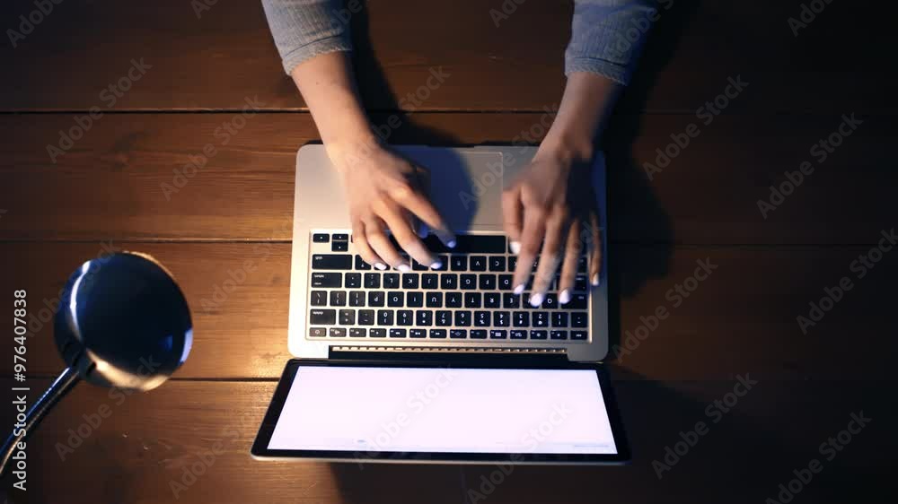 Poster A person is typing on a laptop while using a desk lamp for illumination, creating a cozy work environment in the evening.