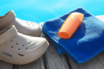 Stylish beach slippers, towel and sunscreen on wooden deck near outdoor swimming pool, closeup