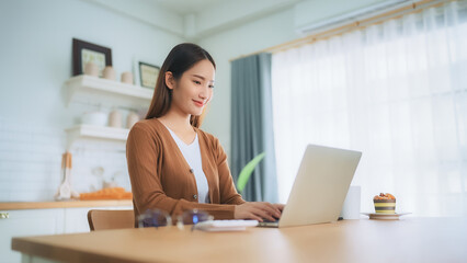 Beautiful young Asian woman working with laptop computer in home kitchen. Work at home