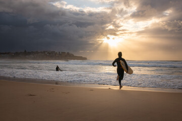 This photo shows a beach scene at sunset with a surfer carrying a board, another in the water,...