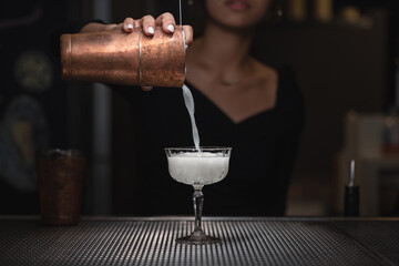 This photo shows a person pouring a white, frothy liquid from a copper shaker into a stemmed glass...
