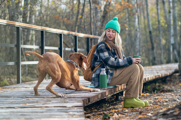 Cheerful hound sticking to resting woman sitting on wooden eco path through thicket drinking tea...