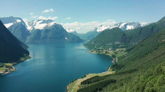 Aerial dolly over forest to deep blue fjord in Norway, surrounded by towering cliffs and mountain ranges with snow capped mountain ridges