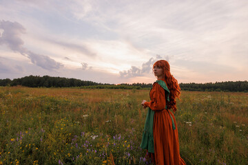 Fototapeta premium Beautiful woman with red hair in long red and green dress standing on flower field. Cloudy sky at sunset. Concept of rural romance, rural scene or love story.