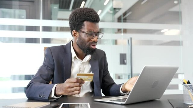 Shocked upset american businessman businessman checks his credit card on laptop while sitting at workplace in business office. Frustrated black man faced fraud or scam. He became a victim of deception