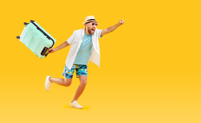 Full body photo of a funny young happy man in sunglasses and beach hat carrying his blue suitcase hurrying up on summer holiday trip on a studio yellow background. Vacation and travel concept.
