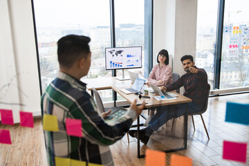 Young professionals engaged in team brainstorming session in modern office. Colleagues discussing ideas using sticky notes and digital data on screen. Creative and collaborative workplace environment