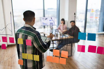 Employeess conducting brainstorming session in modern office with charts on glass wall and colorful...