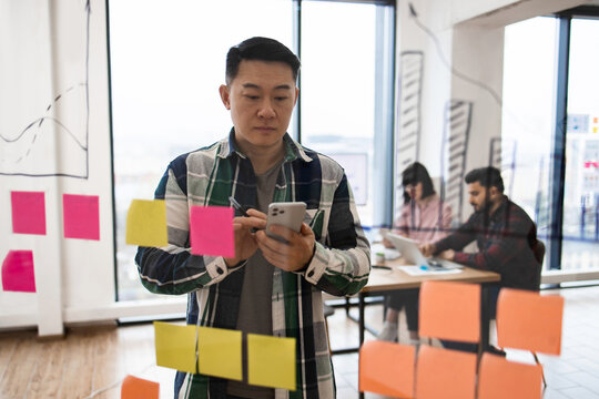 Young professional using smartphone in a modern office environment with brainstorming session and two colleagues working in background. Glass wall, sticky notes, and charts reflect workspace.