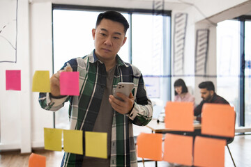 Businessman brainstorming with sticky notes in modern office. Focused man planning with smartphone. Team working together in background.