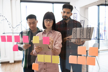 Group of young professionals in collaborative brainstorming session. Team members using sticky notes on glass wall, analyzing data and charts together. Creative and innovative teamwork concep.