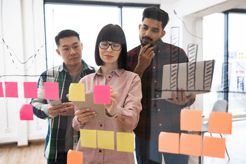 Team of young professionals brainstorming and collaborating using sticky notes on glass wall in modern office. Discussion focused on project planning, organization, and innovation.