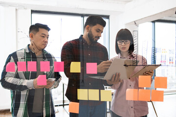 Group of young professionals collaborating on project in modern office. Team gathered in front of glass wall with colorful sticky notes while using digital devices for brainstorming and planning.