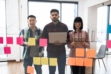 Diverse team collaborating in modern office environment using sticky notes on glass wall. Team members with laptops and tablets planning and brainstorming. Modern workspace with city view.