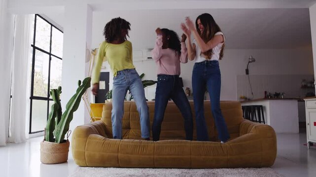 Three young adult female friends having fun together dancing on the sofa at home party. United multiracial women jumping on couch. Friendship and happiness concept.