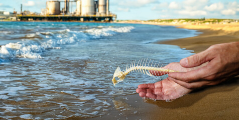 Release of fish skeleton into the sea from the sandy shore. In the blurred background stands a factory plant next to the rough sea.