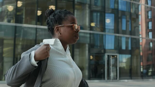 Medium shot of young African American female journalist putting on grey jacket while standing in front of tall glass building in city downtown and preparing for interview or formal recording