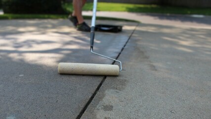 a paint roller for sealing being used on a concrete driveway with a persons legs and paint tray in the background