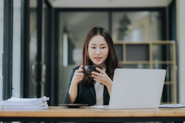 A cheerful young Asian businesswoman sits at her modern desk, typing on a laptop, working on corporate projects with paperwork, smiling in a busy workplace.