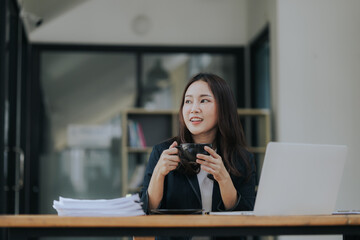 A cheerful young Asian businesswoman sits at her modern desk, typing on a laptop, working on corporate projects with paperwork, smiling in a busy workplace.