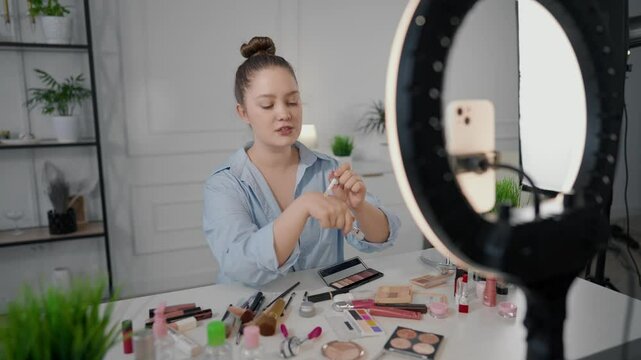 Pretty Caucasian woman showing trendy makeup on her own face, filming tutorial. Beauty blogger streaming in social media by smartphone, view through round lamp, influencer advertising cosmetic brand