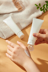 A model gently massages white cream onto her hand, while a second tube is visible behind her, suitable for spa or beauty product advertising. The tube and towel are bathed in sunlight.