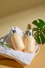 Two cream-colored bottles with pump dispensers are placed on a white towel inside a brown wicker basket. A green leaf is peeking out from the side of the basket. The background features beige surface.